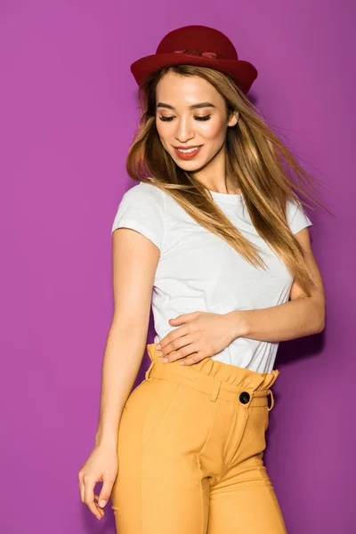 Bella sorridente elegante asiatico ragazza in cappello guardando giù isolato su viola — Foto stock