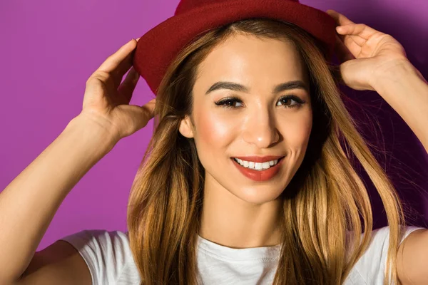 Primer plano retrato de hermosa joven asiática mujer en sombrero sonriendo a cámara aislada en violeta - foto de stock