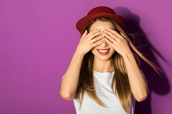 Beautiful cheerful asian girl in hat closing eyes with hands isolated on violet — Stock Photo
