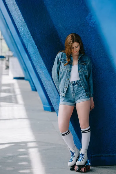 Mujer con estilo en ropa de mezclilla, calcetines altos y patines retro apoyados en la pared azul - foto de stock