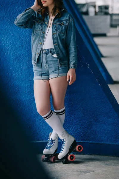 Partial view of fashionable woman in denim clothing and retro roller skates listening music in headphones — Stock Photo