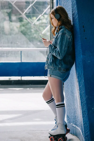 Vue latérale de la femme élégante dans les écouteurs, vêtements en denim, chaussettes hautes et patins à roulettes en utilisant smartphone — Photo de stock