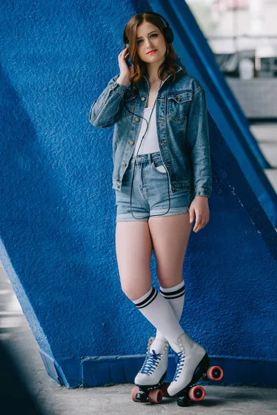 Mujer joven de moda en ropa de mezclilla y patines retro escuchando música en auriculares - foto de stock