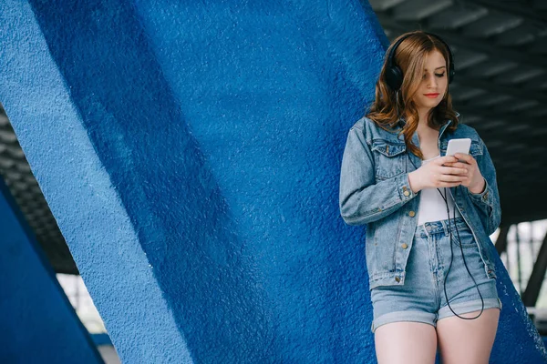 Портрет молодой модной женщины в джинсовой одежде и наушниках с помощью смартфона — стоковое фото