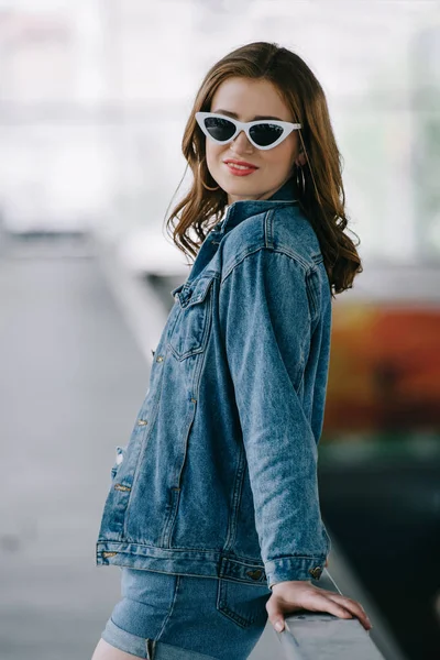 Vue latérale de la femme souriante dans des lunettes de soleil rétro et des vêtements en denim à la mode — Photo de stock