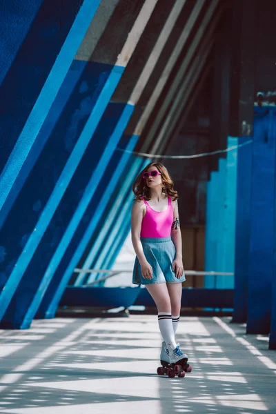 Chica elegante en gafas de sol de color rosa y la falda de mezclilla patinaje sobre ruedas y mirando hacia otro lado - foto de stock