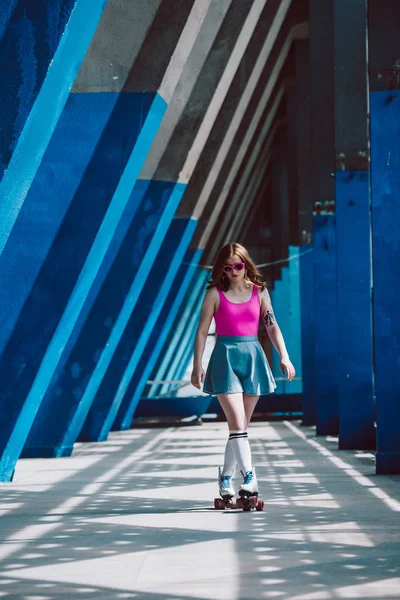 Stylish girl in pink sunglasses and denim skirt roller skating and looking down — Stock Photo