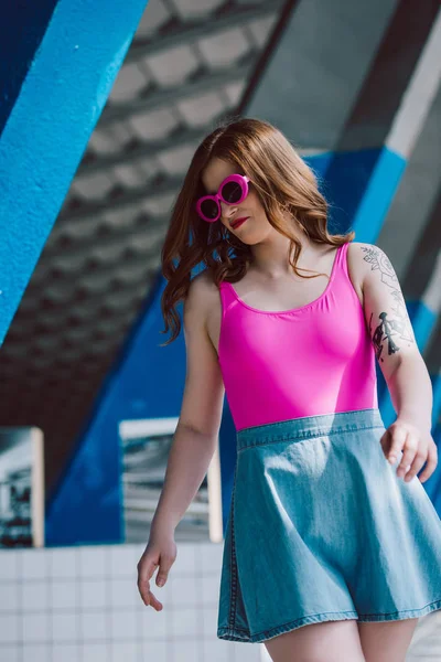 Belle fille élégante en lunettes de soleil roses et jupe en denim regardant vers le bas sur la rue — Photo de stock