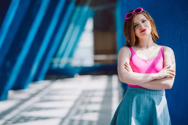 Beautiful young woman in pink sunglasses and denim skirt standing with crossed arms and smiling at camera — Stock Photo