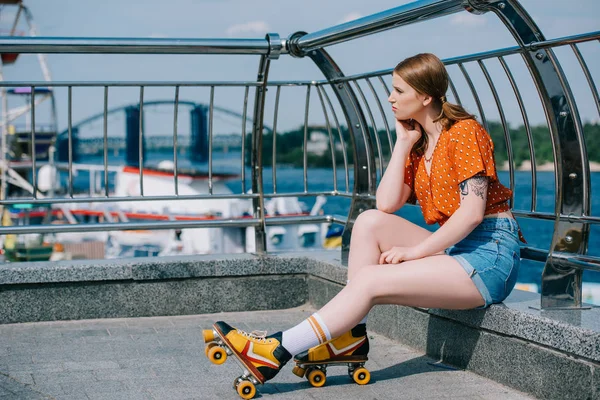 Vista lateral de la chica pensativa en patines mirando hacia otro lado mientras está sentado cerca del río - foto de stock