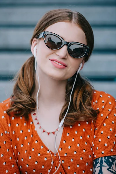 Beautiful young woman in sunglasses smiling at camera while listening music in earphones on street — Stock Photo