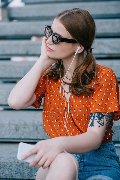 Chica con estilo en gafas de sol y auriculares escuchando música con teléfono inteligente mientras está sentado en las escaleras - foto de stock