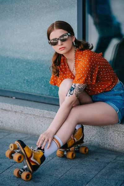 Mädchen mit Sonnenbrille blickt in die Kamera, während sie auf der Straße sitzt und Vintage-Rollschuhe trägt — Stockfoto