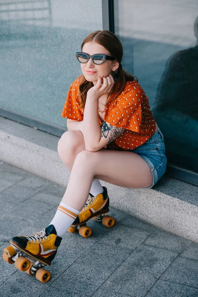 Високий кут зору стильна усміхнена дівчина в сонцезахисних окулярах і роликових ковзанах сидить і дивиться на вулицю — стокове фото