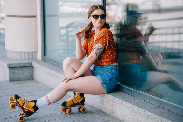 Belle fille en lunettes de soleil et patins à roulettes souriant à la caméra tout en étant assis sur la rue — Photo de stock