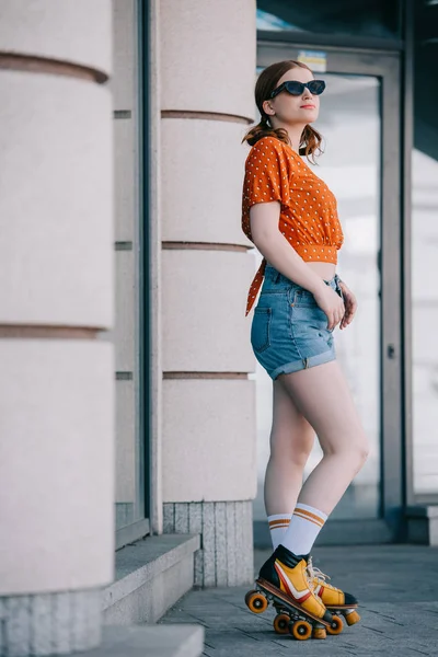 Vista laterale di sorridente ragazza elegante in occhiali da sole e pattini a rotelle in piedi sulla strada — Stock Photo