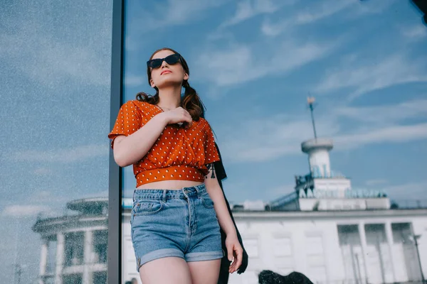 Vista de ángulo bajo de la chica con estilo en gafas de sol y pantalones cortos de mezclilla mirando hacia fuera en la calle - foto de stock