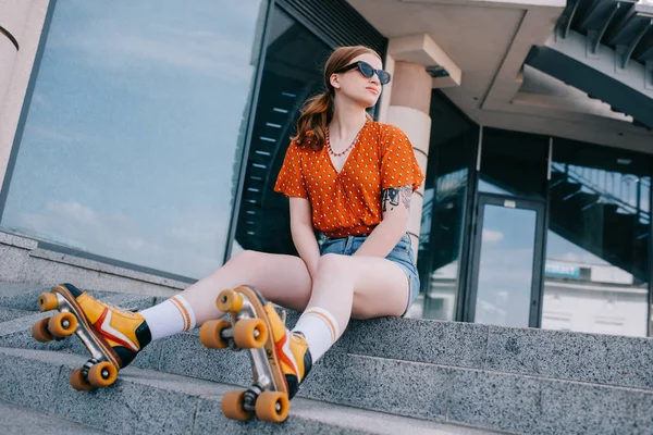Vista de ángulo bajo de chica con estilo en patines de ruedas sentado en las escaleras y mirando hacia otro lado - foto de stock