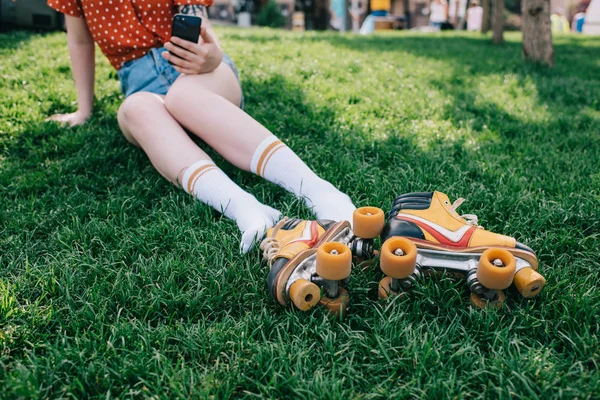 Tiro cortado de menina em meias usando smartphone enquanto descansa na grama com patins — Fotografia de Stock