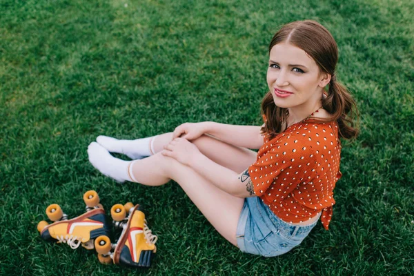 Chica bonita sonriendo a la cámara mientras está sentado en la hierba con patines vintage - foto de stock