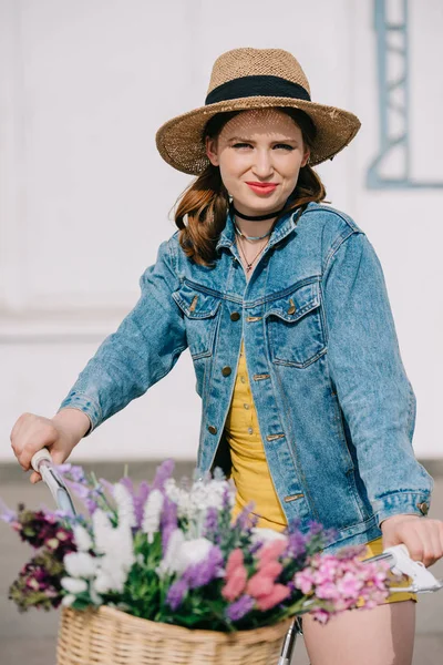 Beautiful young woman in hat and denim jacket riding bicycle and looking at camera — Stock Photo