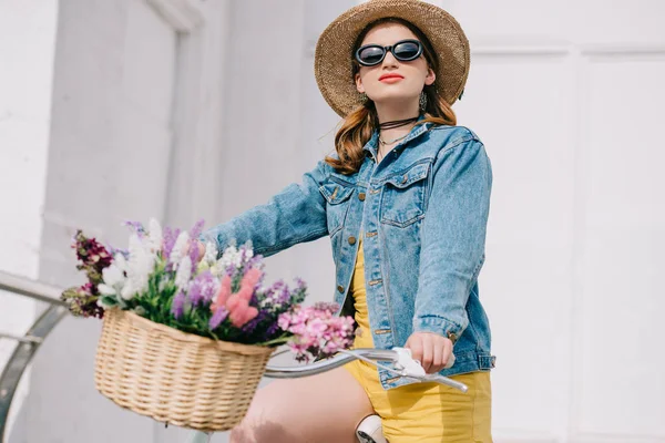 Hermosa chica en sombrero, gafas de sol y chaqueta de mezclilla montar en bicicleta en la calle - foto de stock