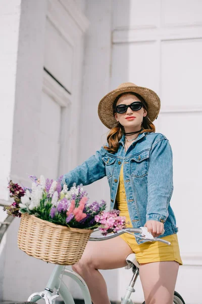 Attraente ragazza in cappello, occhiali da sole e giacca di jeans guardando la fotocamera e in bicicletta sulla strada — Foto stock