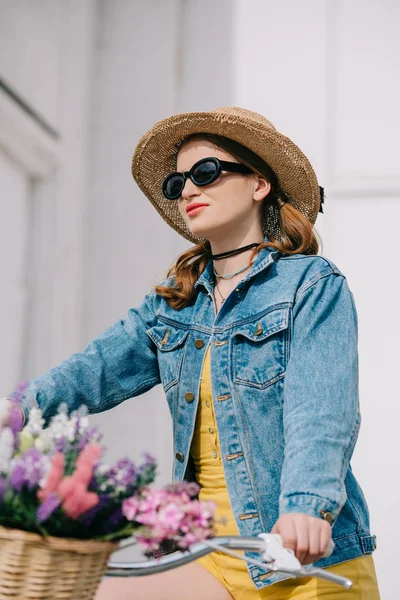 Chica atractiva en sombrero, gafas de sol y chaqueta de mezclilla montar en bicicleta en la calle - foto de stock