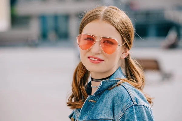 Portrait de belle jeune femme en lunettes de soleil et veste en jean souriant à la caméra — Photo de stock