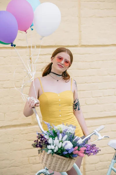 Bela jovem mulher em óculos de sol sorrindo para a câmera enquanto está de pé com bicicleta e balões coloridos na rua — Fotografia de Stock