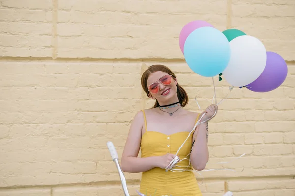 Feliz joven en gafas de sol sonriendo a la cámara y sosteniendo globos mientras está de pie con la bicicleta en la calle - foto de stock