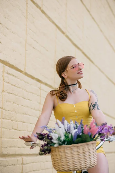 Schönes lächelndes Mädchen mit Sonnenbrille, das beim Fahrradfahren mit Blumenkorb wegschaut — Stockfoto
