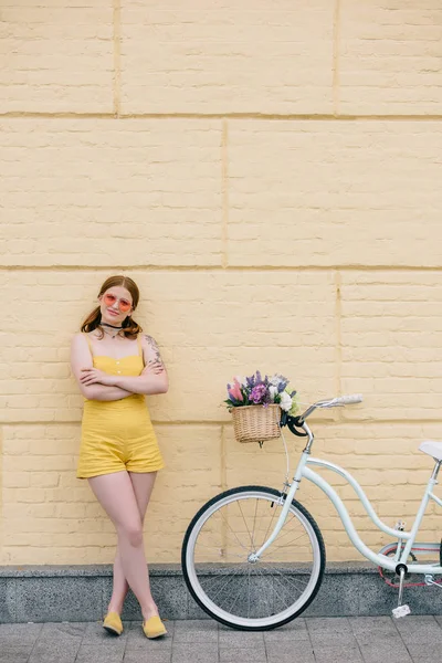 Beautiful young woman in sunglasses standing with crossed arms near bicycle and smiling at camera — Stock Photo