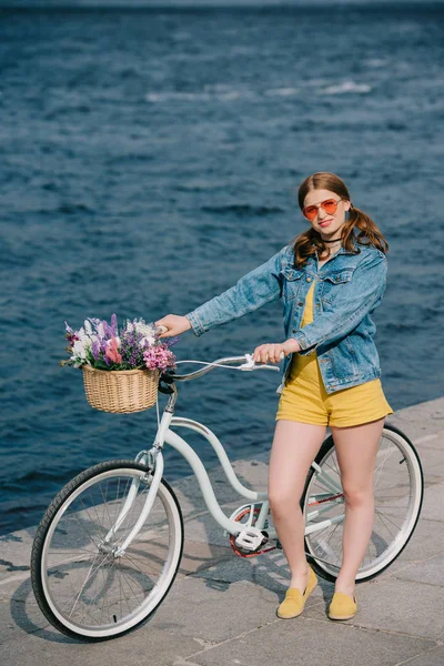 Vista de ángulo alto de la chica en gafas de sol sonriendo a la cámara mientras está de pie con la bicicleta cerca del río - foto de stock