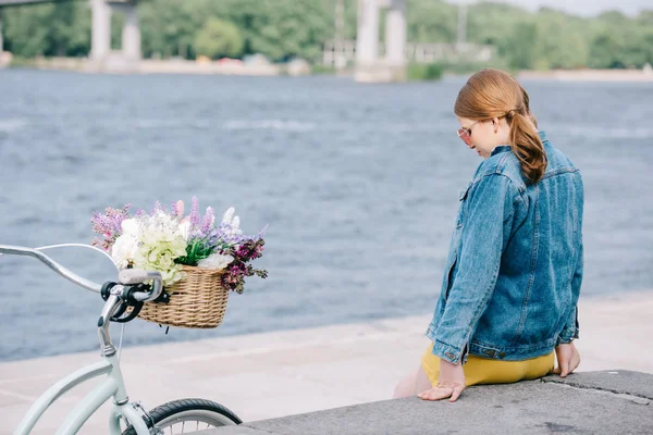 Молодая женщина в солнечных очках сидит на набережной рядом с велосипедом с корзиной цветов — стоковое фото