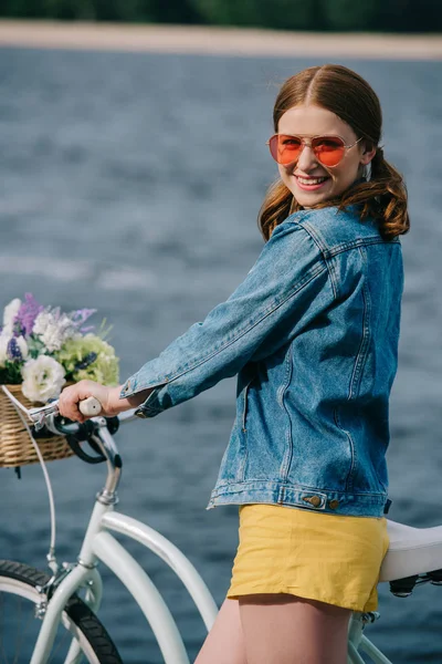 Menina atraente feliz em óculos de sol sorrindo para a câmera enquanto de pé com bicicleta perto do rio — Fotografia de Stock