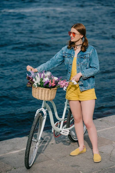 Beautiful stylish girl in sunglasses looking away while standing with bicycle near river — Stock Photo