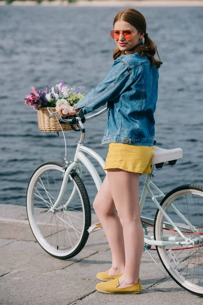 Beautiful young woman in sunglasses smiling at camera while standing with bicycle near river — Stock Photo