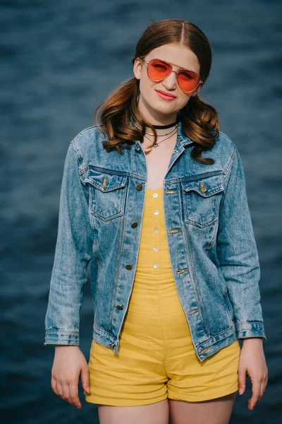 Portrait of beautiful young woman in sunglasses and denim jacket looking at camera outdoors — Stock Photo