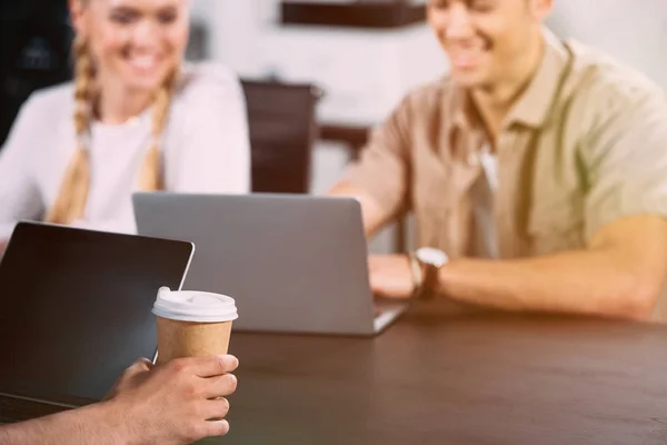 Plan recadré d'un homme d'affaires assis à table avec un ordinateur portable et une tasse de café en papier pendant que ses collègues souriants travaillent derrière — Photo de stock