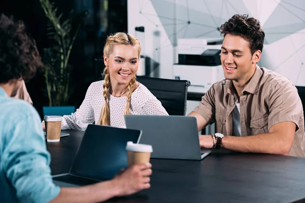 Empresarios sonrientes que trabajan en la mesa con ordenadores portátiles y tazas de café en la oficina moderna - foto de stock