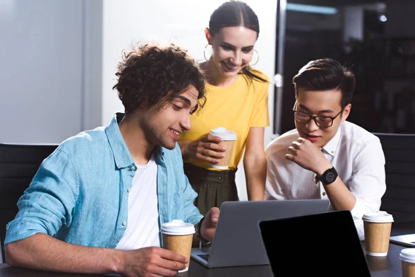 Colegas de negocios multiculturales con tazas de café trabajando y hablando en la mesa con ordenadores portátiles en la oficina moderna - foto de stock