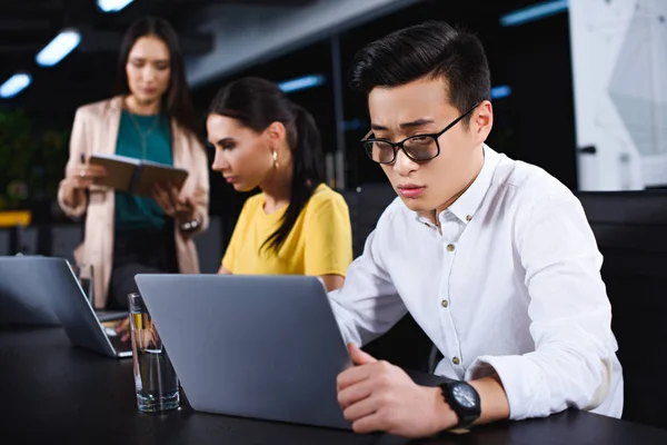 Jovem asiático empresário usando laptop enquanto dois empresárias trabalhando para trás no moderno escritório — Fotografia de Stock