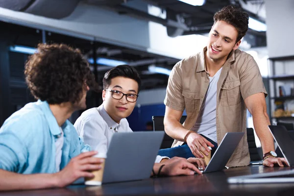 Drei multikulturelle Geschäftsleute treffen sich mit Pappbechern Kaffee am Tisch mit Laptops im modernen Büro — Stockfoto