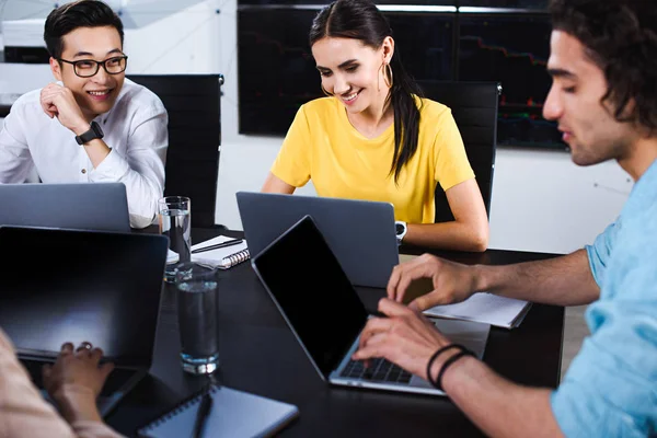 Sorrindo grupo multicultural de parceiro de negócios conversando à mesa com laptops no escritório moderno — Fotografia de Stock