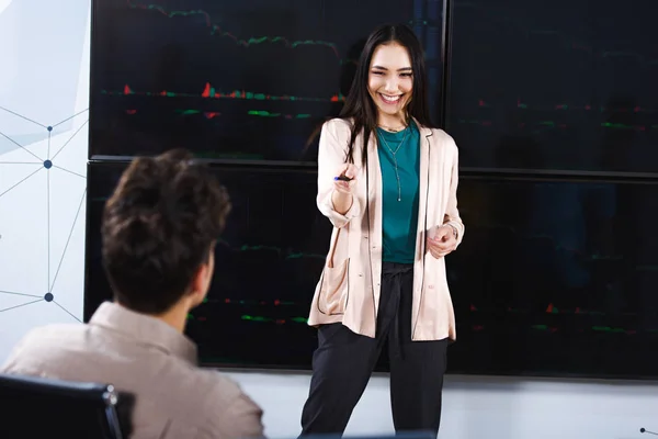 Sonriente asiático mujer de negocios mostrando presentación y apuntando en macho colega en moderno oficina - foto de stock