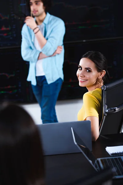 Joven mujer de negocios sonriente mirando a la cámara mientras su socio de negocios muestra la presentación en la oficina moderna - foto de stock