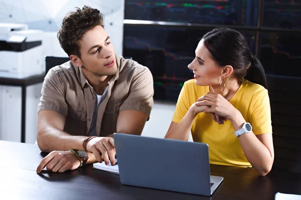 Junger Geschäftsmann zeigt auf Laptop-Bildschirm auf lächelnde Geschäftsfrau am Tisch in modernem Büro — Stockfoto