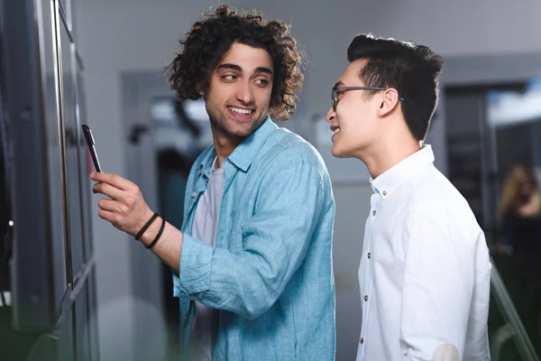 Young businessman pointing by pen at board to asian colleague in modern office — Stock Photo