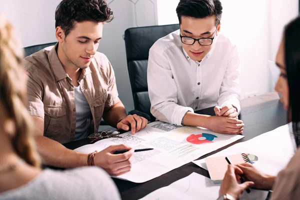 Dos hombres de negocios multiétnicos que se reúnen en la mesa con gráficos en la oficina moderna - foto de stock